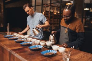 African and caucasian baristas at a wooden counter in a modern roastery, with rows of coffee cups and roasted beans preparing for a tasting